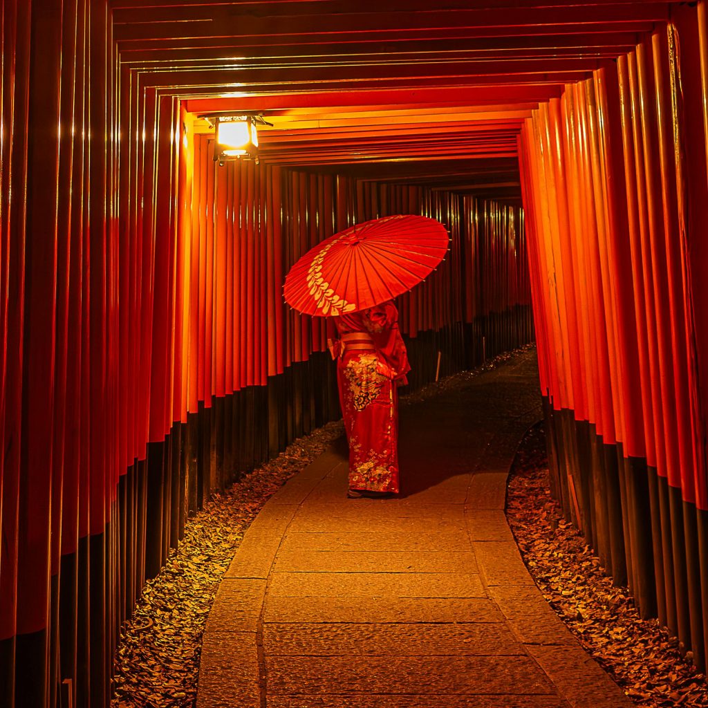 A woman in traditional attire walking with an umbrella under vibrant torii gates at night.