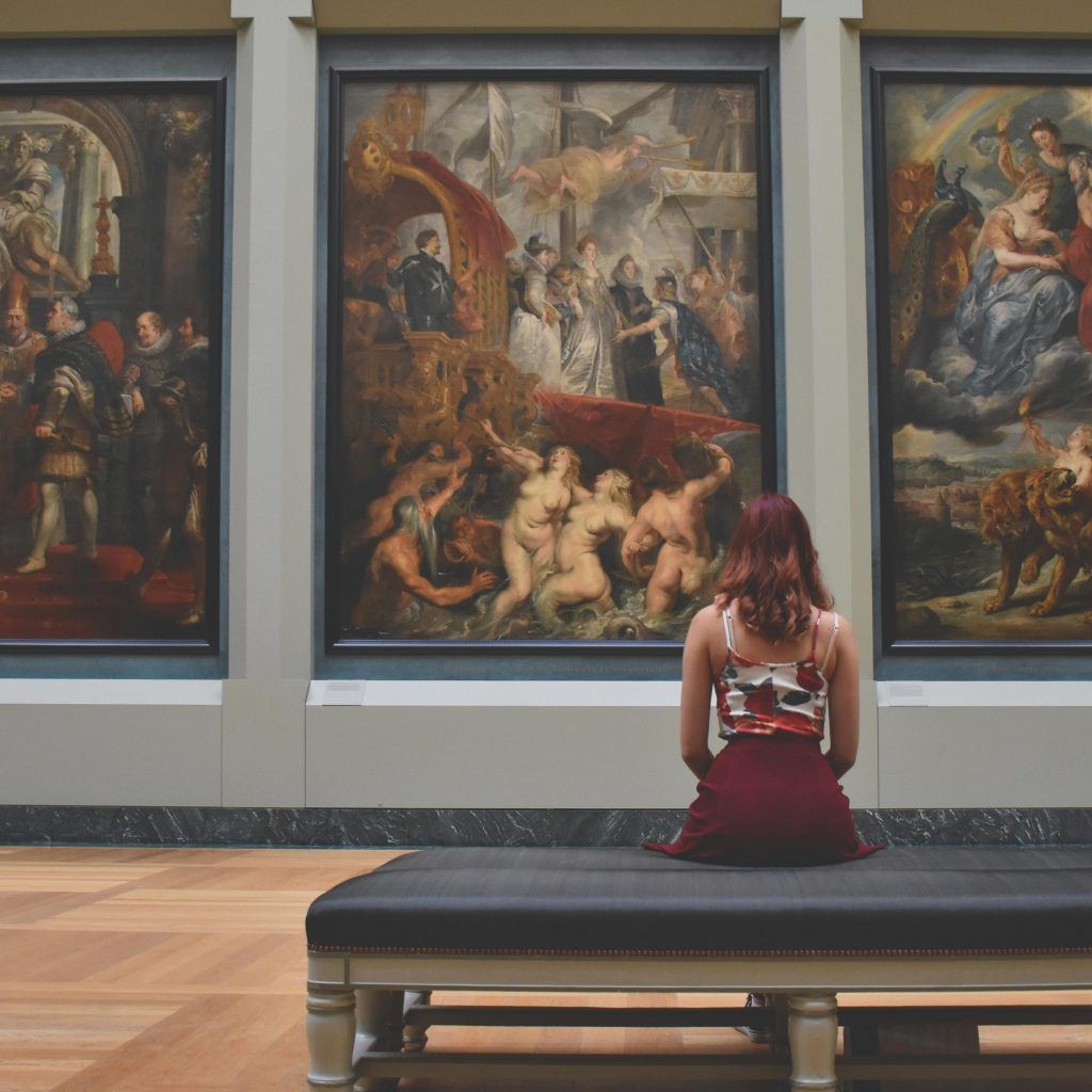 A woman observes classic paintings in an elegant museum gallery.